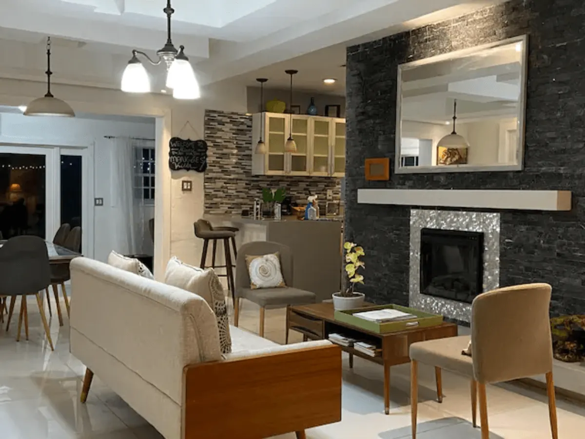 This inviting interior image captures a spacious open floor plan featuring a cozy living area with a sofa and fireplace, a dining table set for a group, and a visible kitchen area in the background. The space is accented by a large mirror and subtle coastal-inspired decor, seamlessly blending comfort and style.