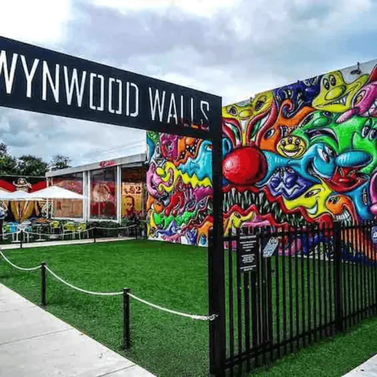 This vibrant exterior shot captures the iconic Wynwood Walls sign, a colorful mural, and an outdoor seating area bustling with energy. The image highlights Miami’s renowned street art scene and the social, community-driven atmosphere of Wynwood, one of the city's most famous entertainment districts.