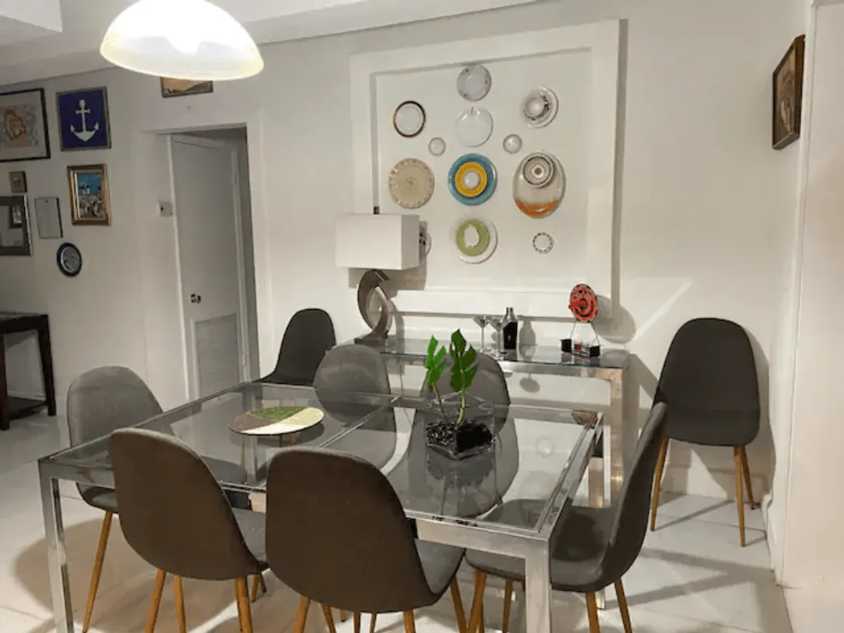 A bright and modern dining area featuring a glass table, contemporary lamp, decorative plate wall, and lush potted plant. The setup invites guests to linger over meals, surrounded by stylish touches that mix mid-century and coastal vibes.
