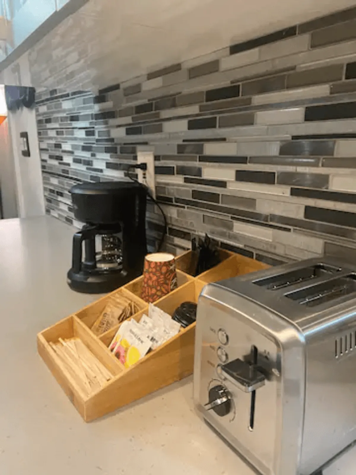 A cozy kitchen setup featuring a coffee maker, toaster, wooden organizer, and a decorative paper cup. The countertop is tidy and functional, highlighting practical amenities for daily routines.