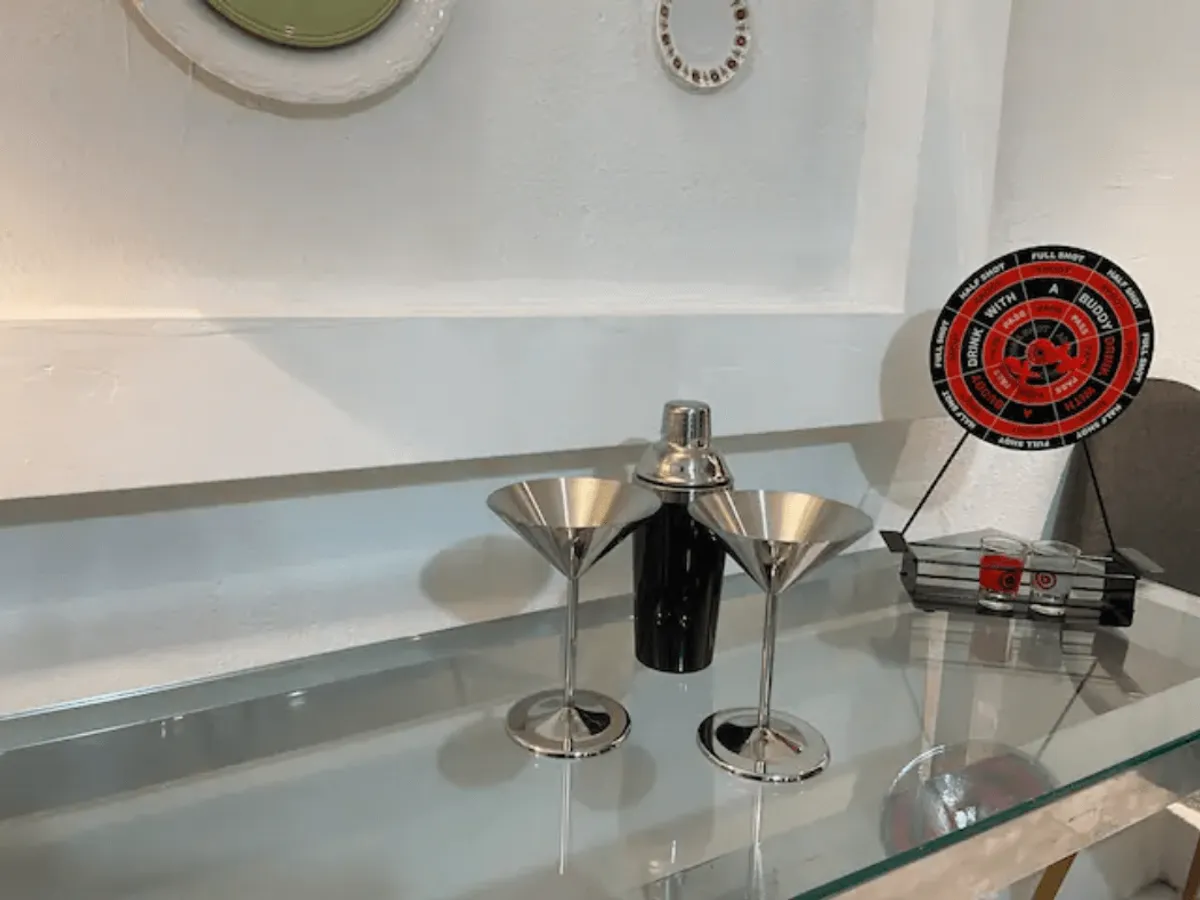 A close-up of two stainless steel martini glasses, a cocktail shaker, and a playful roulette-style drink spinner arranged on a sleek table. The scene hints at mixing cocktails and hosting themed parties with friends.