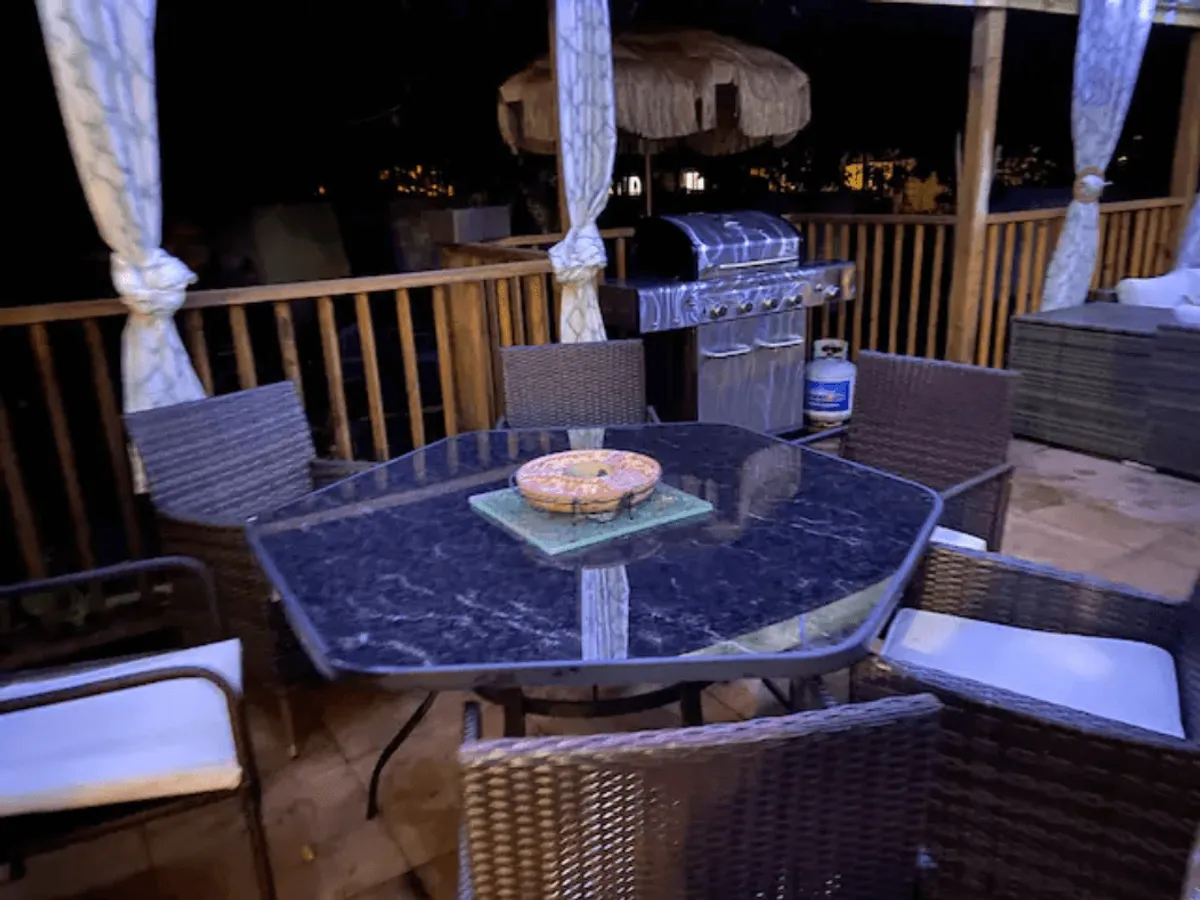 An inviting glass table with woven chairs is arranged for outdoor dining, accompanied by a BBQ grill. The space is designed for al fresco meals and socializing, reinforcing the idea of entertaining and relaxing in a tropical setting.