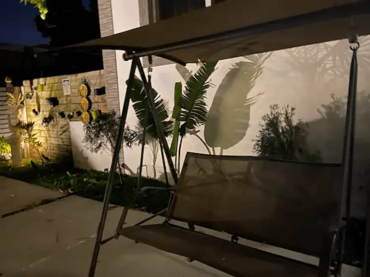 A cozy swing seat under soft outdoor lighting, surrounded by potted plants and plant shadows on an illuminated wall, evoking a serene evening atmosphere ideal for stargazing or peaceful conversation.