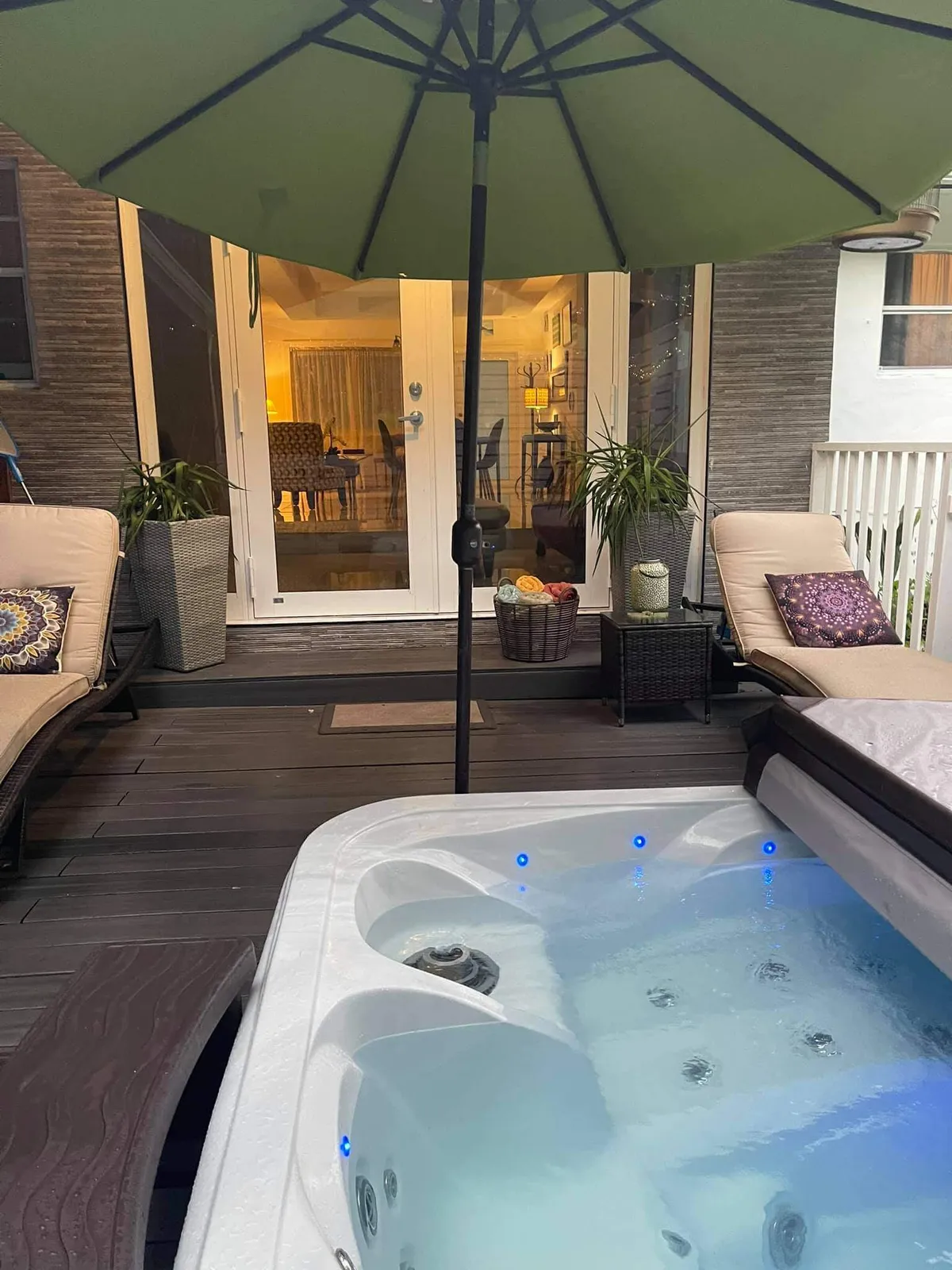 A cozy outdoor area featuring a hot tub illuminated with blue LED lights, two cushioned lounge chairs with patterned pillows, a large green patio umbrella, and glass doors leading into the house. The scene is set for relaxation, with lush landscaping visible around the hot tub and seamless indoor-outdoor flow.