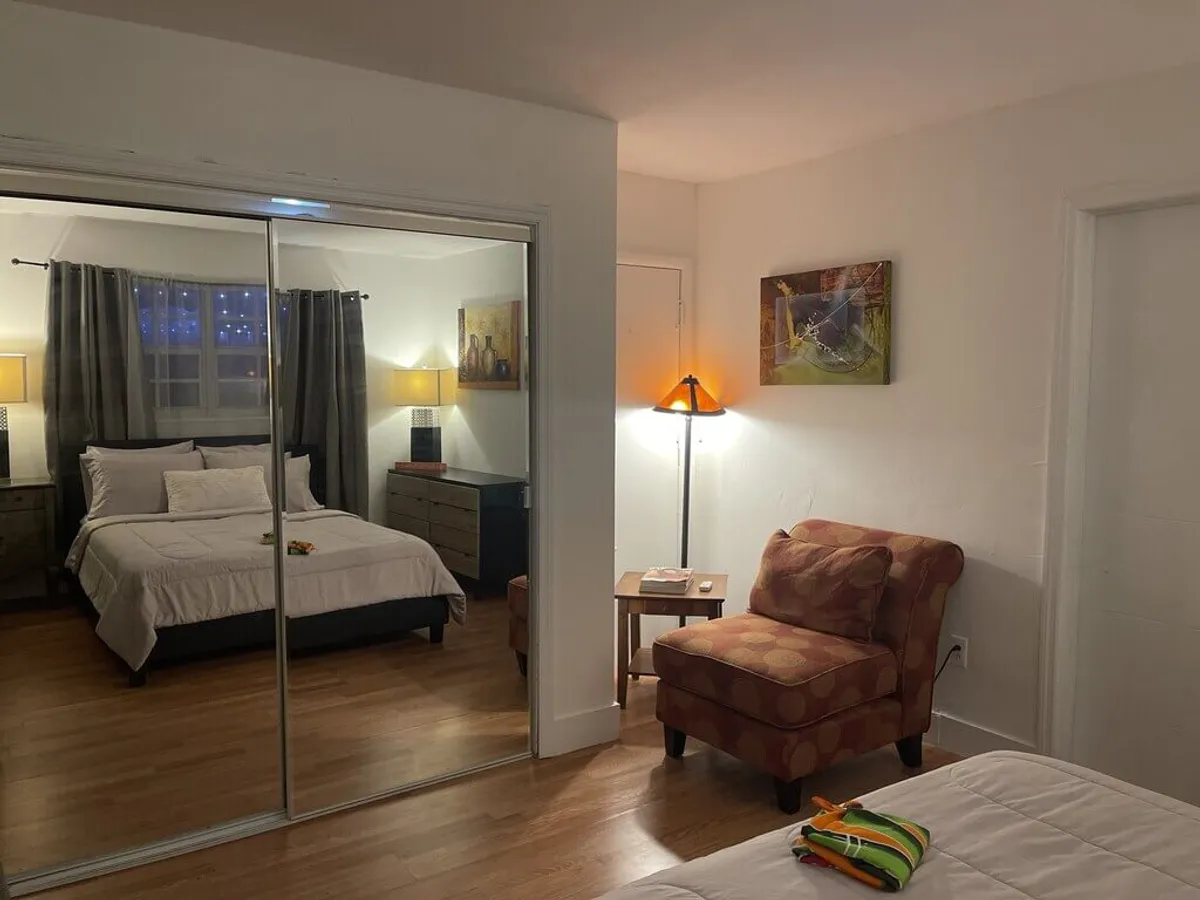 A cozy bedroom featuring a bed with white linens and multiple pillows, mirrored closet doors reflecting the room, a comfortable patterned armchair with a cushion, and a floor lamp casting warm light. The mirrored closet provides significant storage for clothing and gear.