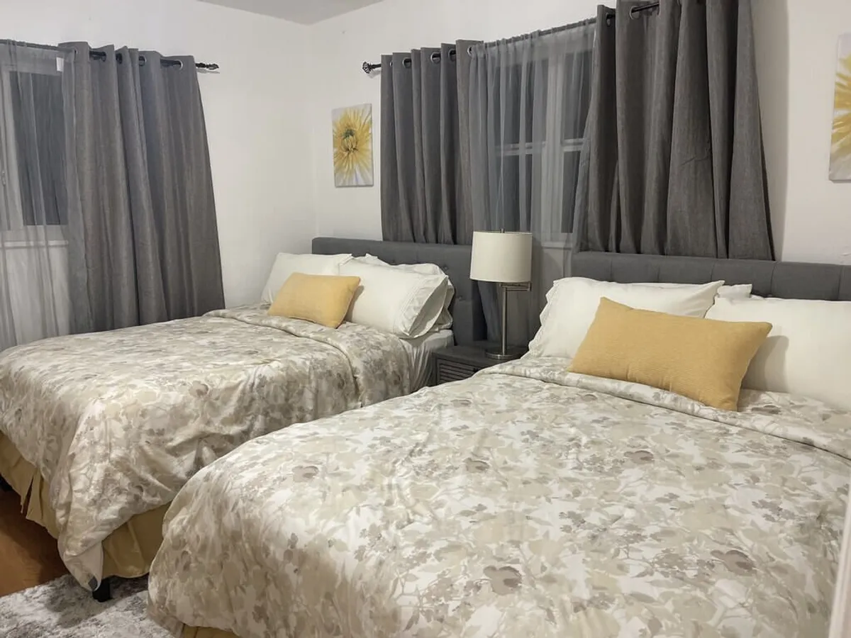 This interior bedroom image features two queen-sized beds with floral patterned bedding, accented by yellow pillows, and grey curtains framing the windows. A nightstand with a lamp sits between the beds, completing the cozy, restful atmosphere.