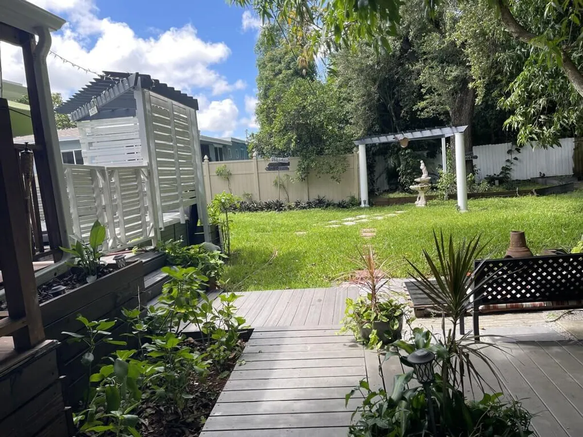A peaceful garden scene with a white pergola structure centered around a birdbath or small fountain, a lush green lawn with mature trees, a wooden deck area with potted plants and a garden bench, and a privacy fence enclosing the yard.