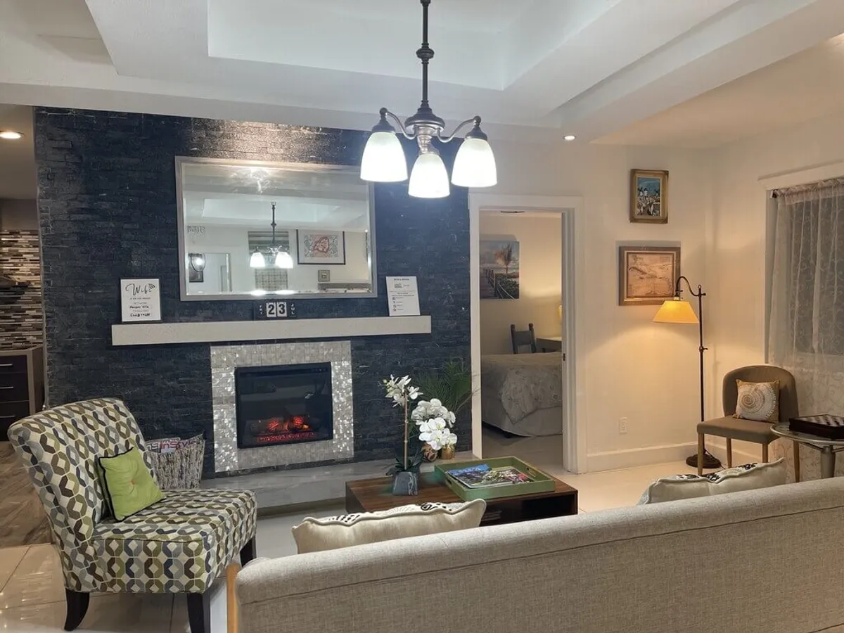 This image showcases a welcoming living area centered around a fireplace with a decorative tile surround and a large mirror above it, reflecting light throughout the space. A patterned accent chair with a green pillow and a coffee table with decorative items and books add cozy, stylish touches. The overall mood is warm, comfortable, and conducive to relaxation or intimate gatherings.