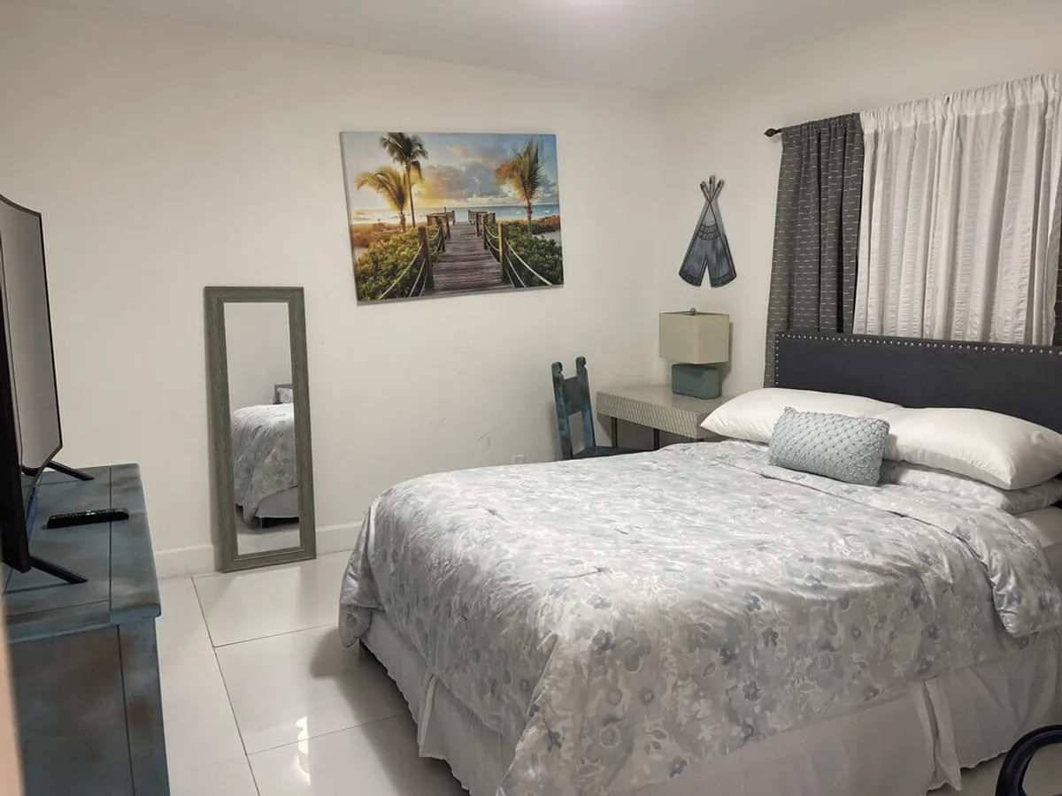 A bedroom with a bed adorned in patterned bedding and pillows, tropical boardwalk wall art, a full-length mirror, and a blue wooden chair with a desk and lamp. The room feels calm, bright, and thoughtfully arranged for both rest and quiet activities.