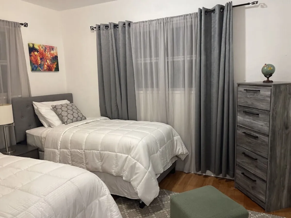 This image captures a bright bedroom with two twin beds dressed in crisp white bedding, a gray dresser topped with a decorative globe, large windows with layered curtains, and floral wall art. The dresser offers visible storage options for clothing and personal belongings.