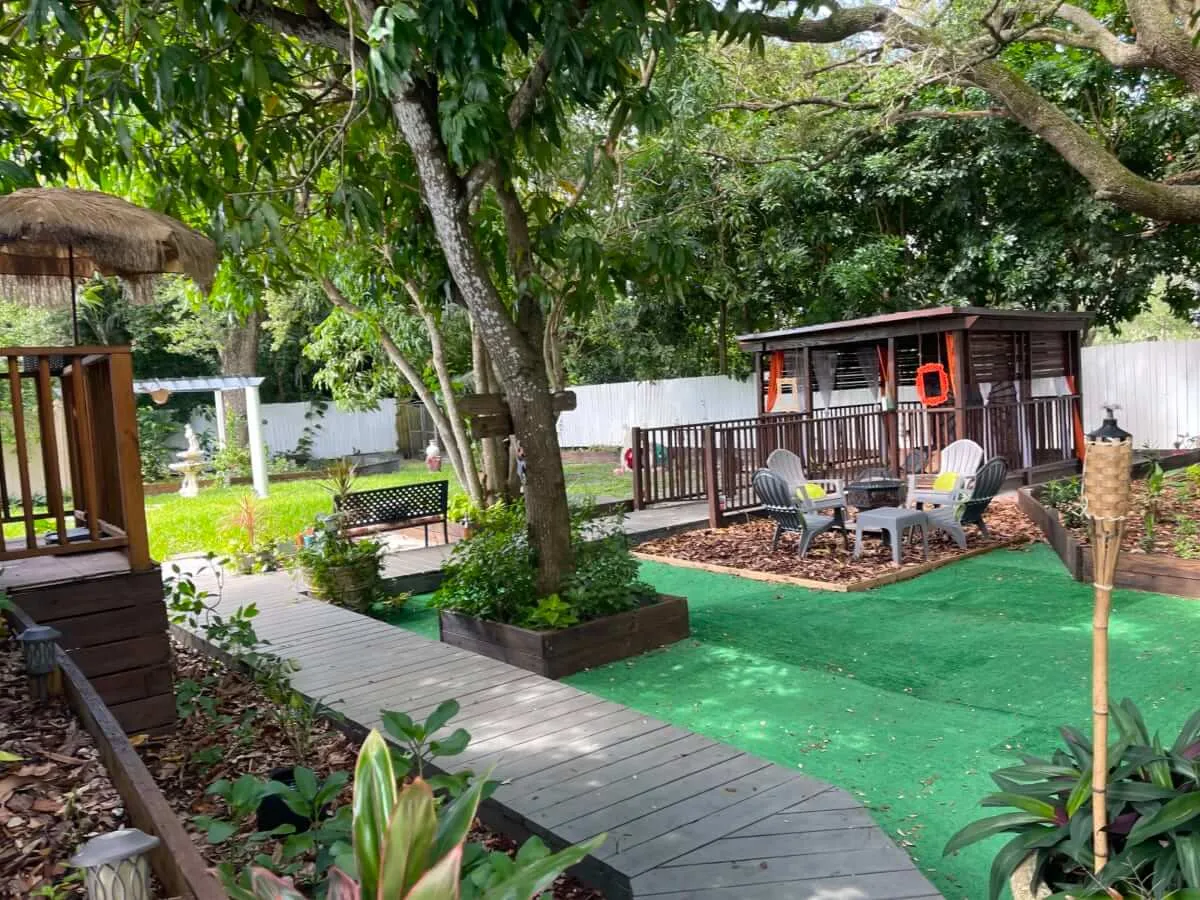 A charming chickee hut with a thatched roof, surrounded by a seating area featuring Adirondack chairs and a table. Wooden walkways weave through garden beds and mature trees provide ample shade, creating a tranquil retreat for gatherings or quiet moments outdoors.