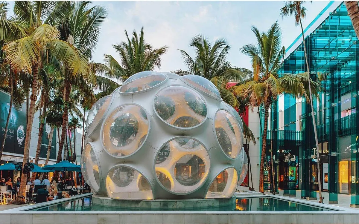 A striking exterior shot of a large spherical art installation surrounded by a shallow reflective pool, with tall palm trees and modern glass storefronts lining the area. People can be seen walking, shopping, and socializing outdoors, hinting at a lively atmosphere. The setting seamlessly blends urban energy with tropical elements, capturing the spirit of Miami’s Design District.